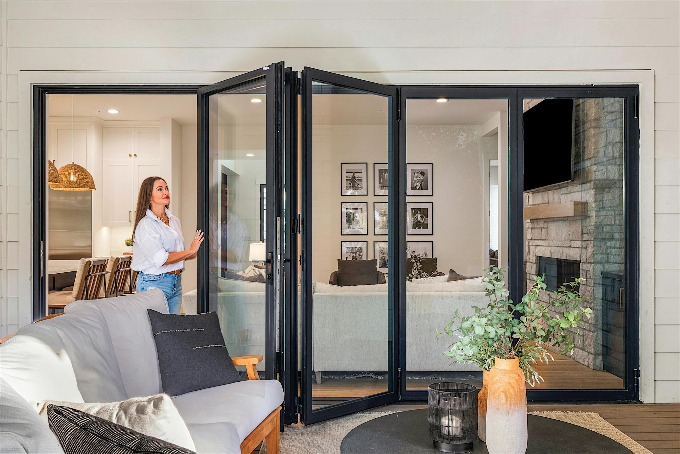 Molly Mesnick opening expansive bifold glass doors to a living room featuring a gray sofa, black coffee table, and decorative plants.